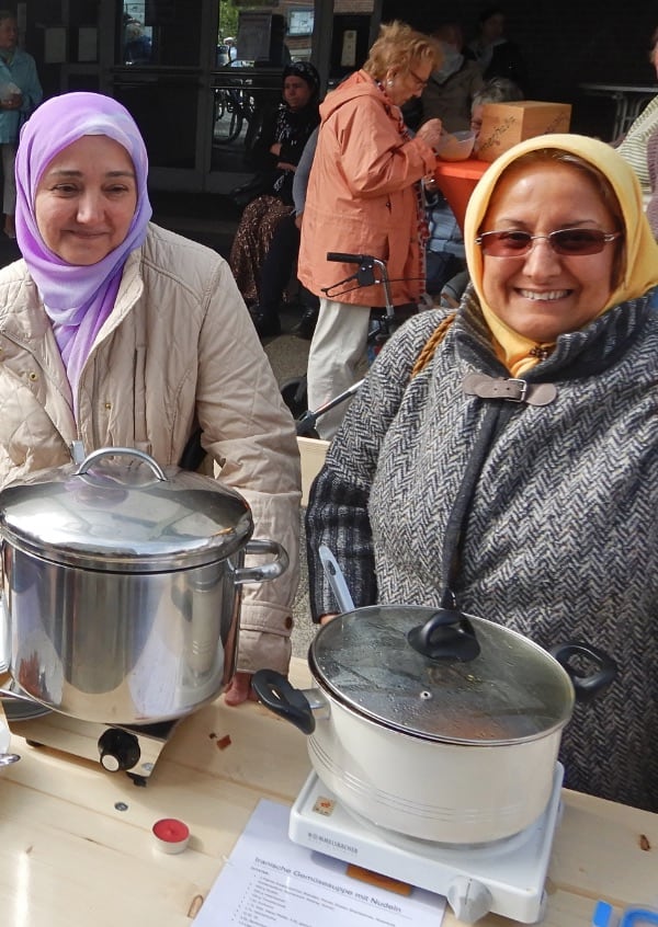 Zwei lächelnde Menschen mit Kopftuch, die im Rahmen einer Veranstaltung ihre selbstgemachte Iranische Gemüsesuppe anbieten
